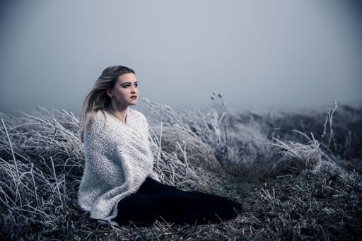A cute blond girl wearing a knitted scarf sitting in a field covered in frost looking aside
