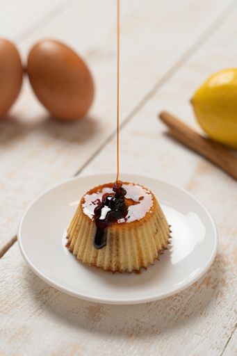A selective focus closeup of a plate of Creme Caramel dessert, cinnamon, lemon, and eggs on the white plank table