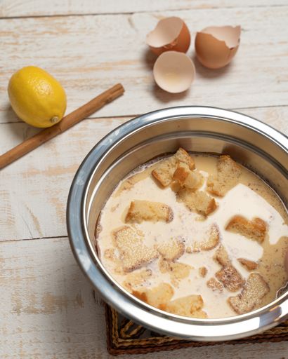 A vertical shot of a bowl of milk with bread, cracked eggshells, lemon, and cinnamon on a plank table