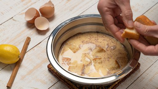 A closeup of a hand putting bread on a bowl of milk, cracked eggshells, lemon, and cinnamon on a plank table