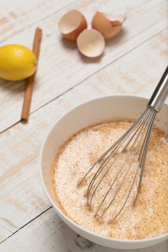 A vertical closeup of a bowl of mixed ingredients with an egg beater, cracked eggshells, lemon, and cinnamon on a plank table