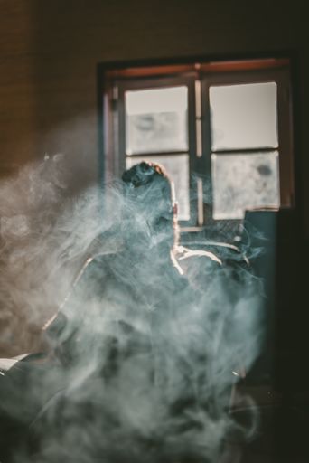 A vertical shot of a person sitting on a chair and smoking filling the room with smoke - smoking danger concept