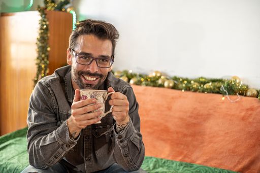 A shallow focus of a smiling handsome young Caucasian man in glasses sitting on a sofa at home with a cup of tea at Christmas time