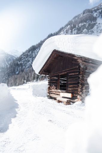 A vertical shot of a wooden house covered in snow with mountains in the background on a sunny day