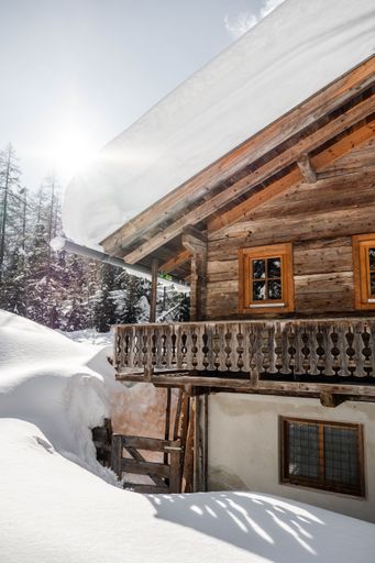 A vertical shot of a wooden house covered in snow with mountains in the background on a sunny day