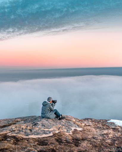 A vertical shot of a male sitting on the top of the rock and photographing the foggy cloudy sky