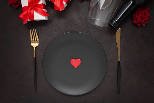 A closeup of a dinner plate, knife, fork, wine bottle, wine glass, gifts, and roses on a black background
