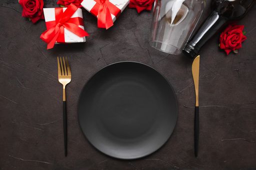 A closeup of a dinner plate, knife, fork, wine bottle, wine glass, gifts, and roses on a black background