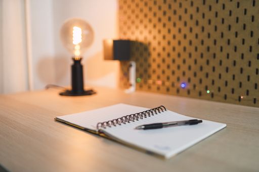 A selective focus shot of an open notebook with a black pen and a lamp on a wooden desk