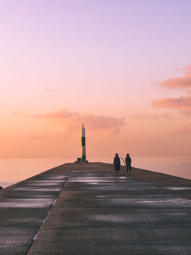 A beautiful nature scene with a sidewalk on the beach with walking people and a lighthouse at sunset