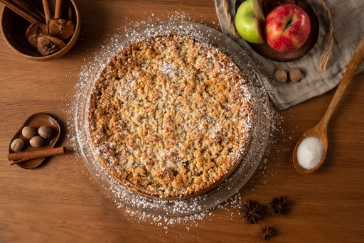 A top view of an apple crumble pie on a wooden table with cinnamon and apples in the background