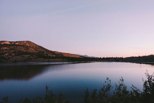 A scenic shot of a still lake surrounded by a hillside and plants at a calm sunset
