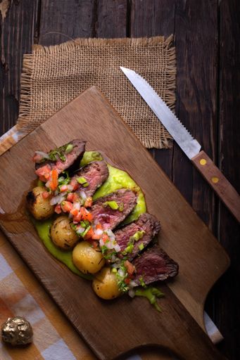 A vertical shot of beef steaks with roasted potatoes, sauce, and vegetable salad on a wooden board