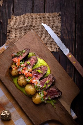 A vertical shot of beef steaks with roasted potatoes, sauce, and vegetable salad on a wooden board