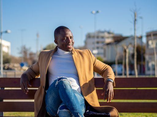 A shallow focus of an adult stylish African male wearing jeans with a turtleneck and beige coat and sitting on a bench