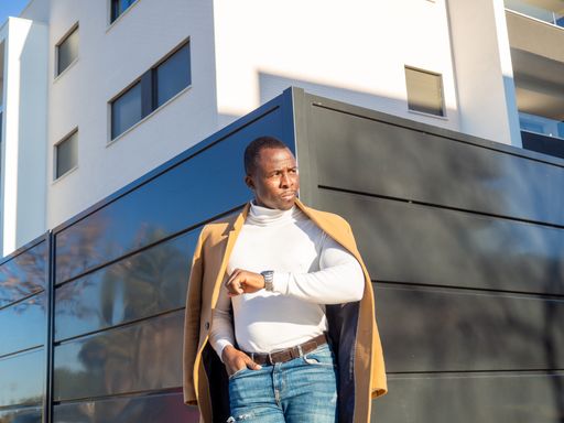 A stylish African male showing his watch on the wrist while leaning on a wall outdoors