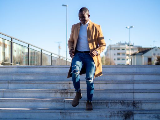 A shallow focus of an adult African male wearing jeans with a white turtleneck and beige coat and walking down the stairs