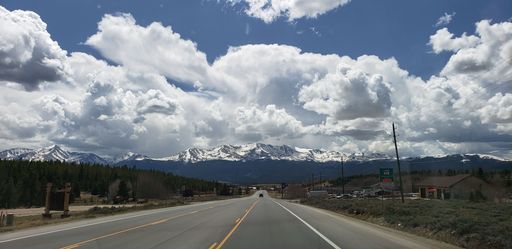 A panoramic shot of the road between the fir forest trees and mountains against a blue clouded sky