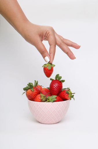 A vertical shot of a hand picking strawberries from a pink bowl on a white surface