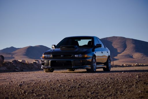 A 1995 Subaru Impreza sedan with turbo custom wheels on a backdrop of blue sky, mountains, and desert landscape