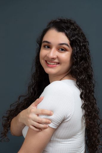 Cheerful beautiful young woman with hand on shoulder wearing white shirt. Against gray background. concept of youth