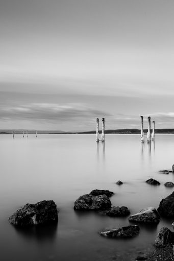 A vertical greyscale shot of a wooden pier near rock formations under the cloudy sky