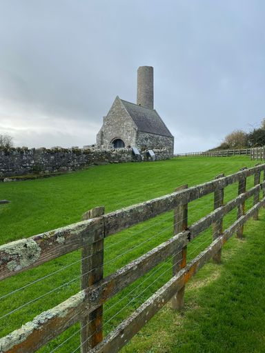 A rural landscape featuring a stone house in the background, surrounded by a white fence and lush green grass in the foreground.