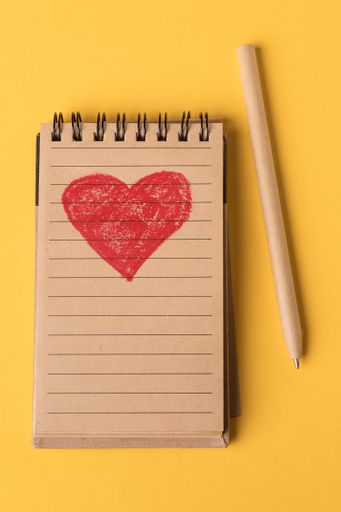 Close-up of a red heart painted in a recycled paper notebook and pen on a yellow background. Top view.