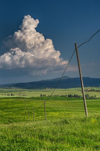 A vertical shot of a green field with hanging wires in val d'orcia tuscany italy with mountains and cloudy blue sky in the background