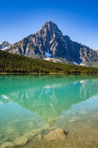 A magnificent view of a transparent mountain lake's surface, with an evergreen forest and snowy mountain slopes in the background, on a sunny day