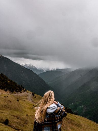 A scenic vertical shot of a blonde woman enjoying the view of evergreen mountains on a gloomy day