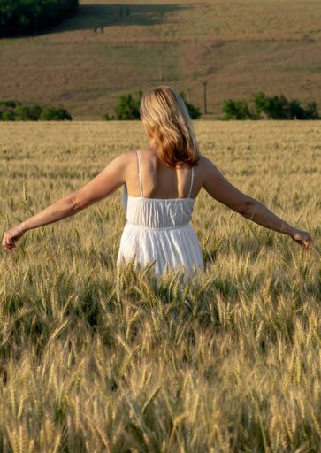 A vertical shot of a young woman in a white dress standing in the barley field during the golden hour