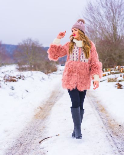 A vertical shot of a female wearing a pink fur jacket, pants, winter boots, and a hat in the snow