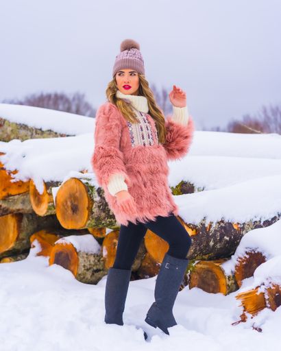 A vertical shot of a female wearing a pink fur jacket, pants, winter boots, and a purple hat in the snow