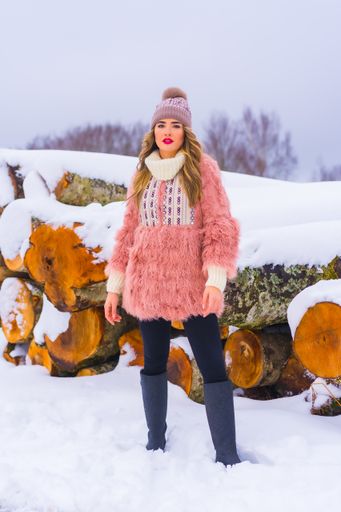 A vertical shot of a female wearing a pink fur jacket, pants, winter boots, and a purple hat in the snow