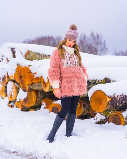 A vertical shot of a female wearing a pink fur jacket, pants, winter boots, and a purple hat in the snow