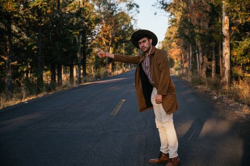A young Hispanic man with cap and coat showing thumb up while hitchhiking on the road