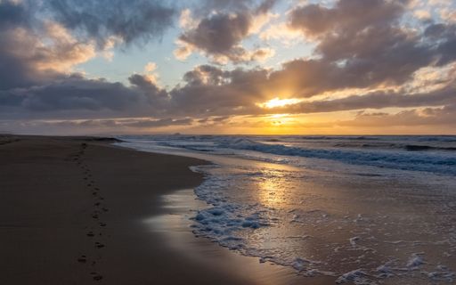 A scenic beach landscape with turbulent waves crashing against the shore against the cloudy sunset sky