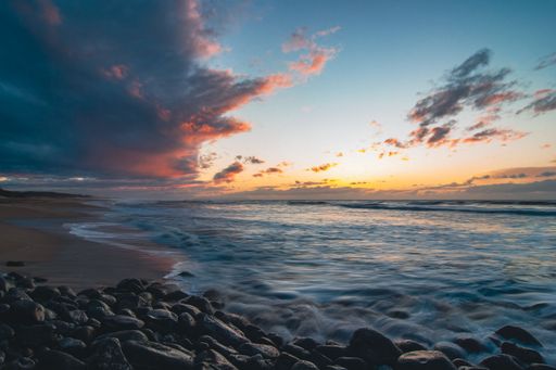 A scenic beach landscape with turbulent waves crashing against the shore against the cloudy sunset sky
