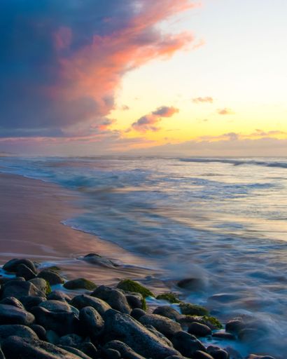 A scenic beach landscape with turbulent waves crashing against the shore against the cloudy sunset sky