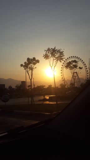 A vertical shot of a brightly shining sunset sun over a ferris wheel in a park seen from a car window