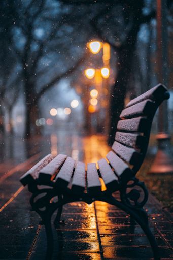 The snow beautifully falling on a wooden bench covered in snow in the park, with bokeh lights behind it