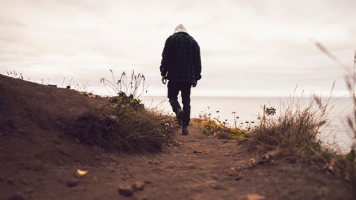 An adult male with a hoodie walking along a sandy path by the shore on a cloudy day