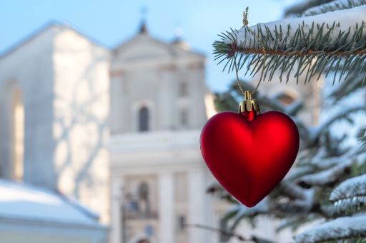 A cute and festive red heart ornament hanging from a tree branch against a backdrop of a church