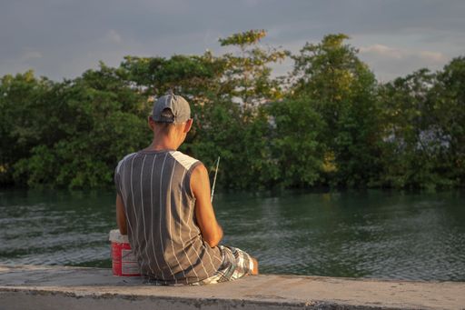 A male fisherman stands on the bank of the River San Juan de Matanzas in Cuba, proudly holding a fishing rod