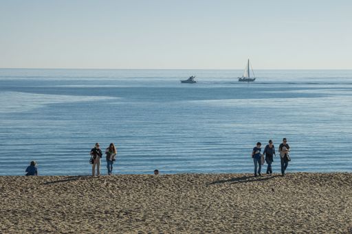 A group of people, at the sandy beach in Barcelona, Spain, enjoying the sunshine, with a boats sailing on the sea in the background