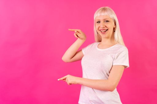 A portrait of a blonde caucasian woman pointing with her fingers in front of a pink background