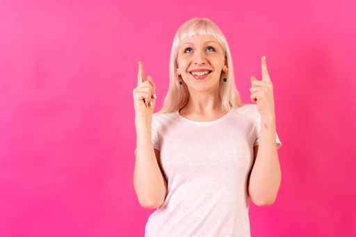 A portrait of a blonde caucasian woman pointing with her fingers in front of a pink background