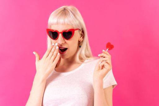 A portrait of a blonde caucasian woman wearing heart-shaped glasses in front of a pink background