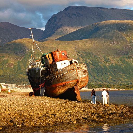 A group of people inspecting an old wrecked wooden ship on a rocky shore surrounded by tall green hills